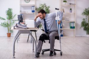 A man with a crutch at a desk. he is wincing in pain and his knee is in a brace. 