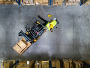 We see an aerial view of a forklift accident in a warehouse. An injured man lays on the floor. 