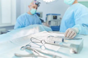 A surgeon going to pick up a tool from a tray of surgical equipment with another medical professional in the background.