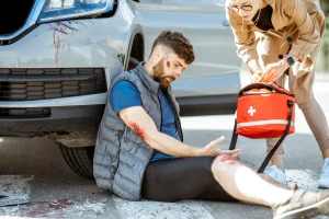 A driver sits against his care while receiving first aid for multiple injuries