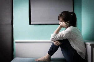 A woman sits alone on a bed with the blinds shut. 