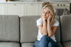 A woman is sitting on a sofa with a pained face and her eyes shut. 